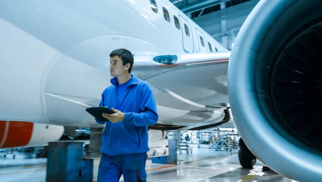 Aircraft maintenance trainee holding a tablet beside an airliner engine in a hangar