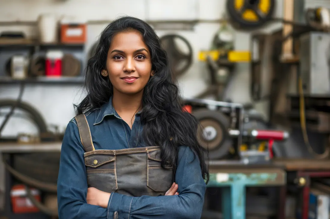 Female mechanic in a denim work apron standing with arms crossed in an engineering workshop