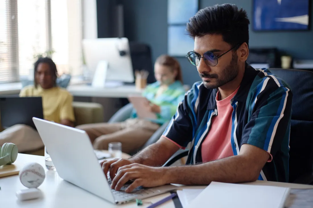 Indian man working on a laptop at a shared desk, preparing for aircraft maintenance module exams