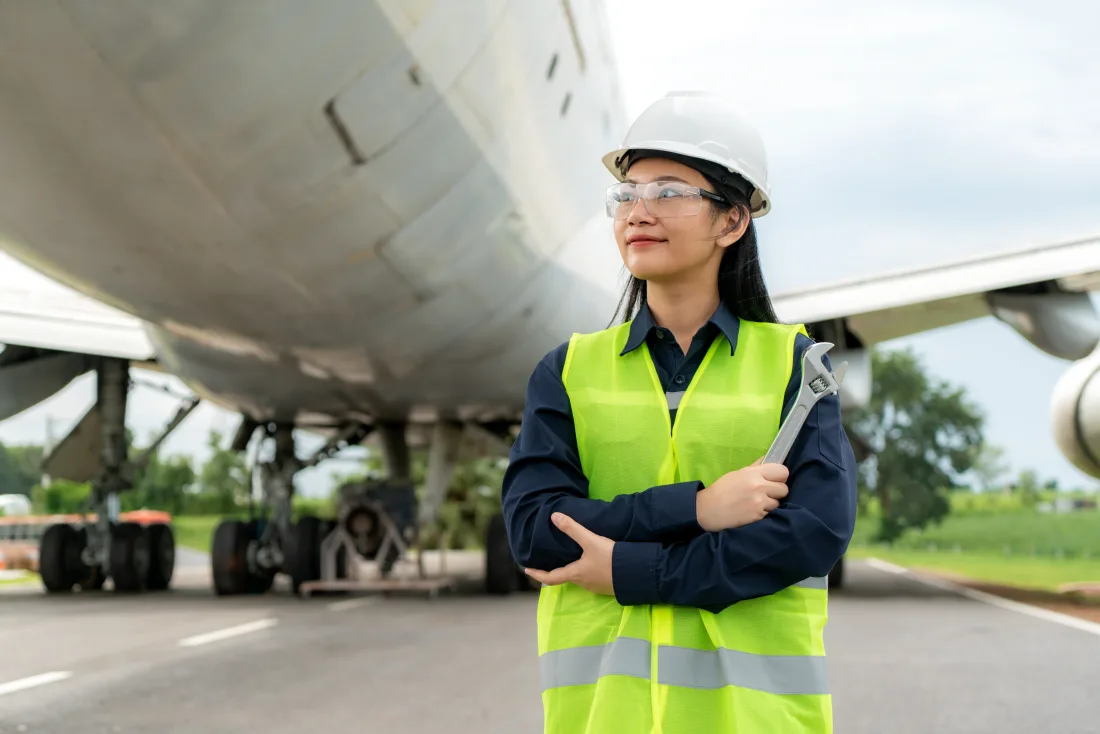 Woman engineer in a hi-vis vest and white helmet standing beside an airliner on the apron