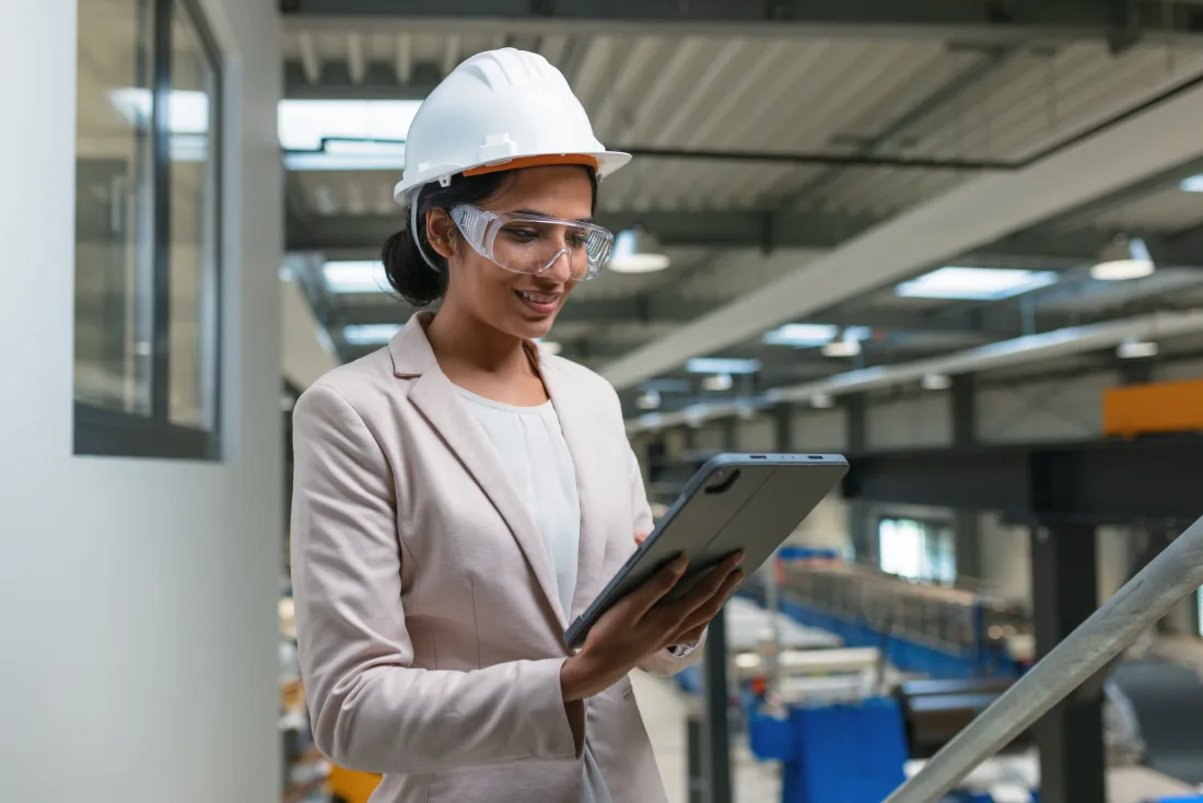 Woman in a white safety helmet and safety glasses checking a tablet in an industrial facility
